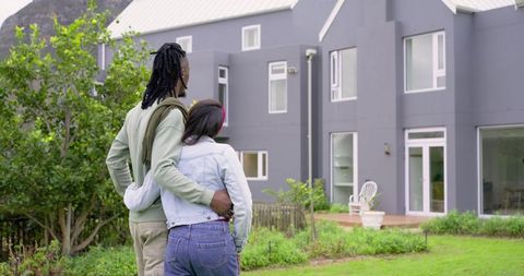 Diverse young couple embracing and looking at modern grey house with garden and lawn
