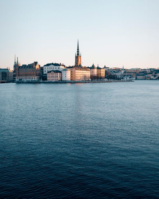 Photograph of Stockholm's waterfront at sunset showing historic buildings and reflecting light on calm water. Useful for travel guides, tourism ads, cultural magazines, or postcards.