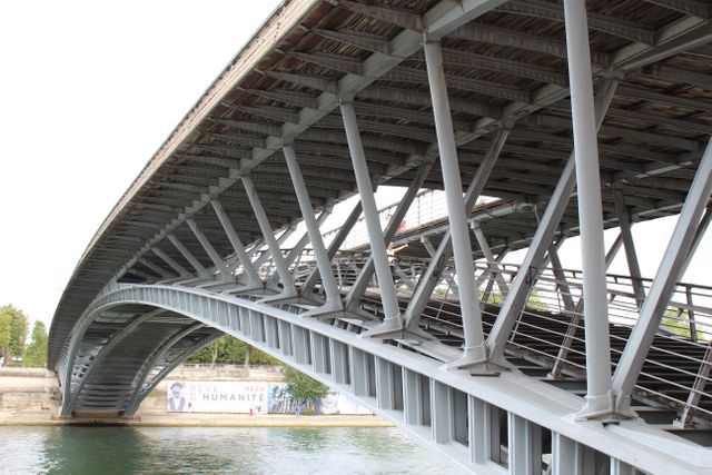 This image depicts a modern steel bridge spanning over a calm body of water in an urban landscape. The architecture emphasizes industrial design and structural engineering. This photo can be used in projects related to civil engineering, urban development, modern infrastructure, or architectural magazines. It is also ideal for website headers, travel blogs, and promotional material for city tours.