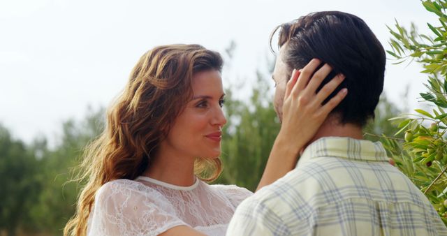 A Caucasian woman and man share a tender moment outdoors, with the woman gently touching the man's face, with copy space. Their affectionate gaze suggests a romantic connection in a serene natural setting.
