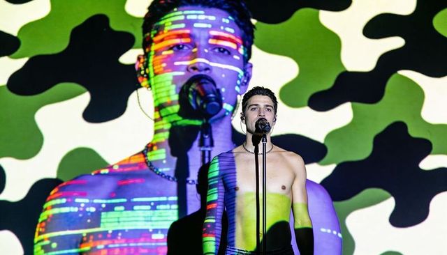 Shirtless singer performing with glitch camouflage projections on concert stage spotlight