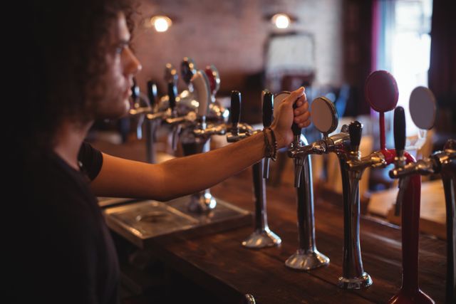 Male bartender operating beer taps at pub counter. Ideal for use in articles about nightlife, hospitality industry, bar culture, and beverage service. Suitable for illustrating concepts related to bartending, pubs, and social gatherings.