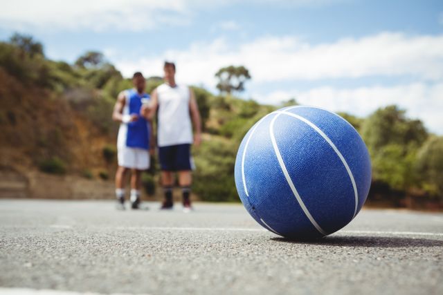 Blue basketball lying on an outdoor court with two players standing in the background. Ideal for use in sports-related content, fitness promotions, teamwork concepts, and recreational activity advertisements.