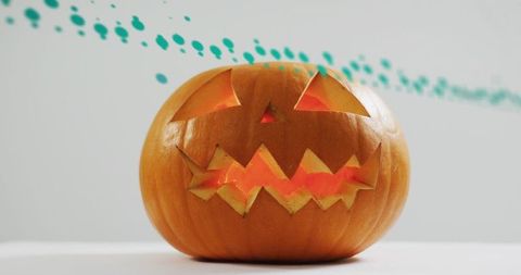 Glowing carved pumpkin casting warm amber light on white tabletop with turquoise paint streak