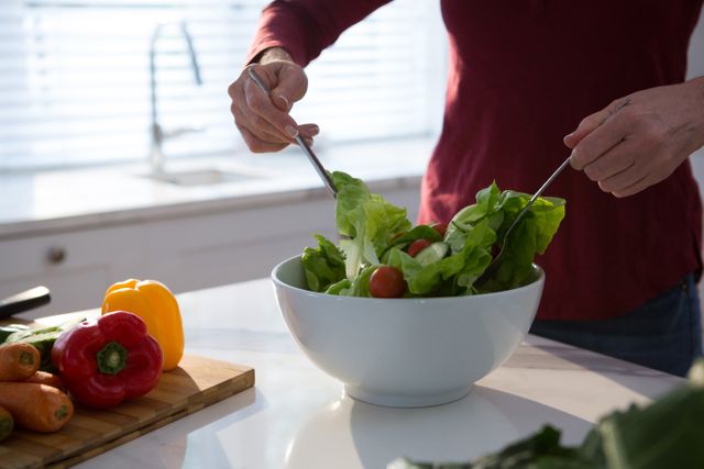 Mid section of woman preparing a fresh salad in a modern kitchen. Ideal for use in articles or advertisements related to healthy eating, cooking at home, vegetarian recipes, wellness, and lifestyle blogs. The image highlights fresh ingredients and a clean, bright kitchen environment.