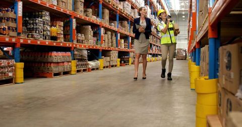 Two women walking warehouse aisle inspecting inventory with tablet and handheld scanner