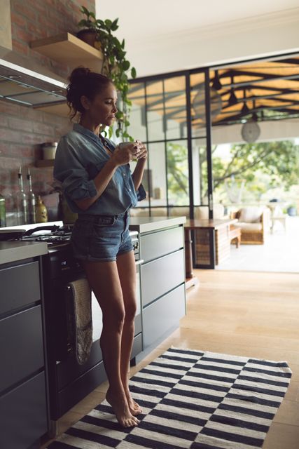 Woman standing in a modern kitchen, drinking coffee and looking thoughtful. She is wearing casual denim shorts and a shirt, barefoot on a striped rug. The kitchen features sleek cabinets, indoor plants, and large windows letting in natural light. Ideal for use in lifestyle blogs, home decor magazines, and advertisements promoting comfortable living spaces.