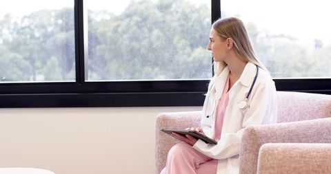Female clinician sitting in pink armchair looking out window holding tablet and stethoscope