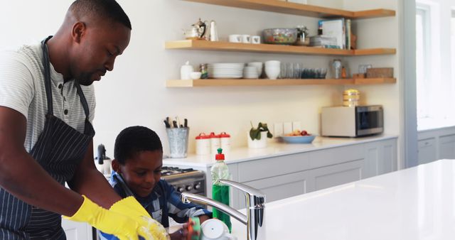 An African American middle-aged man and a boy are washing dishes together in a bright kitchen, with copy space. They share a moment of domestic life, highlighting the importance of family participation in household chores.