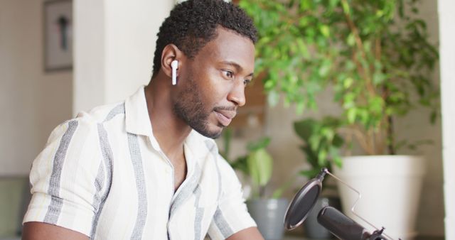 Young man wearing airpods, leaning towards a professional microphone in a home office environment, focusing on content creation or podcasting. Well-suited for media related to communication, technology, home office setups, modern workforce, and digital content creation.