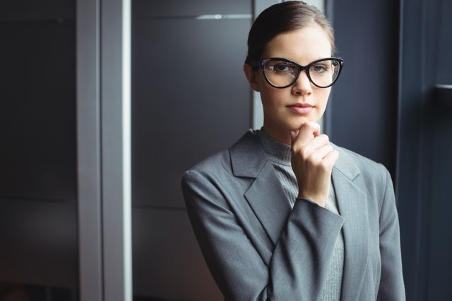 This image depicts a confident female counselor in a modern office setting, wearing glasses and business attire. She is thoughtfully holding her chin, suggesting deep thinking or decision-making. This image can be used for business, corporate, or professional themes, including articles on leadership, counseling, executive coaching, and workplace environments.