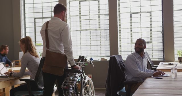 A busy modern office with professionals engaging in different tasks. One man with a messenger bag is beside his bicycle engaging with his seated colleague. Other workers are focusing on their laptops, showcasing teamwork and collaboration. Ideal for depicting startup culture, modern work environments, and professional collaboration.