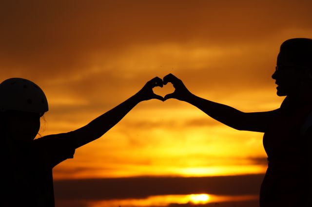 Two people, backlit by warm sunset light, creating a heart shape with their hands. Ideal for concepts like love, friendship, connection, and emotional bonds. Suitable for greeting cards, social media posts, romantic or inspirational content, and lifestyle advertising.