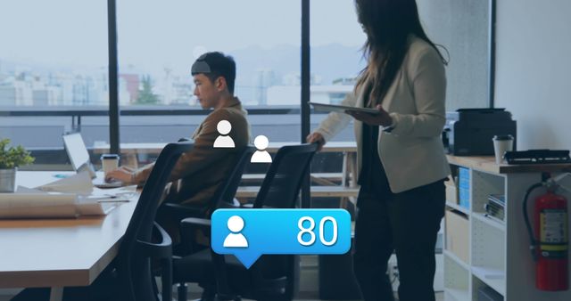 Professionals working in a modern office space with focus on social media connectivity. A woman uses a tablet while a man works on a laptop. Floating icons indicate social media interaction. Suitable for themes on workplace technology, digital collaboration, and social media marketing.