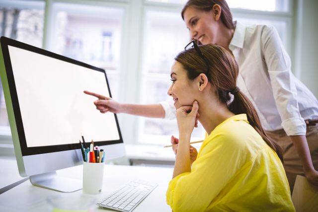 Two women collaborating in a modern office, with one pointing at a computer screen while the other looks on and smiles. Ideal for use in articles or advertisements about teamwork, office environments, professional collaboration, and creative workspaces.