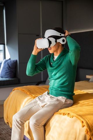 Young man adjusting virtual reality headset while sitting on bed with mustard throw