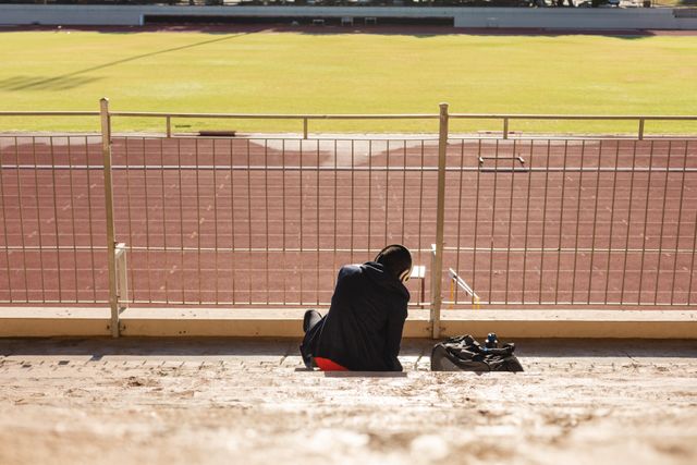 Rear view of fit, biracial male athlete at an outdoor sports stadium, preparing for workout in the stands wearing headphones. Athletics sport training.