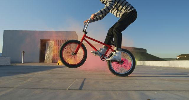 Side view close up of a young Caucasian man riding a BMX bike and doing tricks on the rooftop of an abandoned warehouse, with a pink smoke grenade attached to the bike and colourful smoke grenades in the background