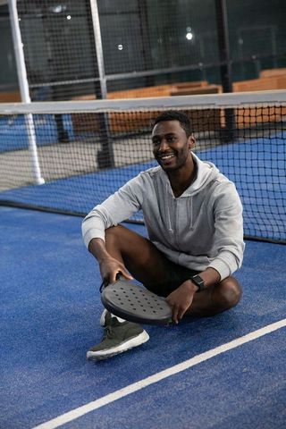 Smiling african american padel player sitting on blue court holding racket in hoodie