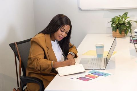 African american businesswoman writing in notebook at modern minimalist desk with laptop