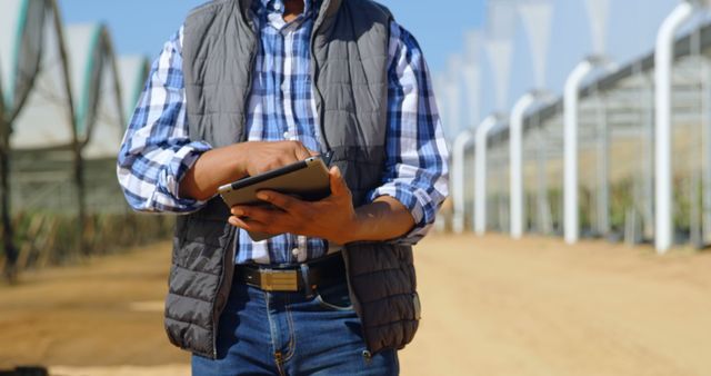 Man standing in modern greenhouse, using tablet for monitoring crops and conducting field analysis. Suitable for illustrating smart farming, digital transformation in agriculture, or precision farming techniques.