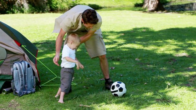 Father and young son enjoying a sunny day in a park, playing and bonding over football. Ideal for use in advertisements promoting family time, outdoor activities, or parenting content. Can also be used in magazine articles about childhood experiences, parent-child relationships, or recreational activities.