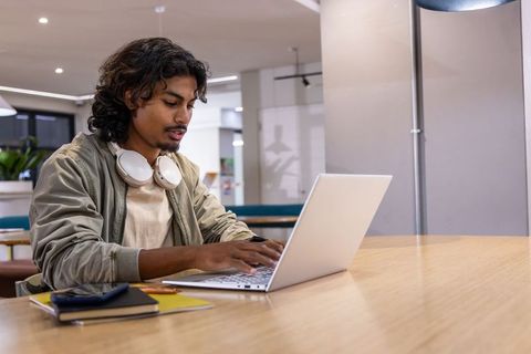 Asian man mid-20s working on laptop in coworking space wearing headphones, khaki jacket