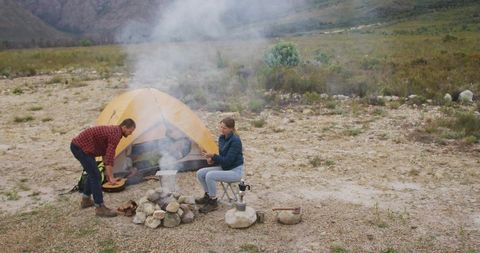 Couple cooking over campfire beside yellow dome tent in remote rocky wilderness landscape