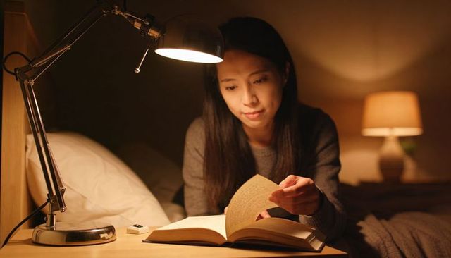 Asian woman reading in bed under warm lamp, cozy nighttime bedtime reading