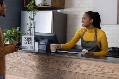 Smiling african american barista handing coffee to customer paying with phone at cafe