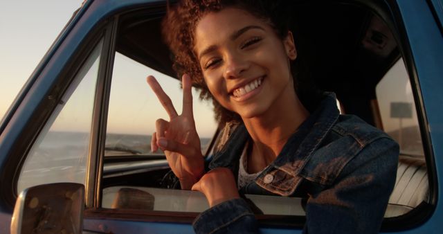 Young woman with curly hair enjoying a road trip. She is leaning out of an open car window, smiling and showing a peace sign with her hand. This image can be used for travel promotions, road trip advertisements, lifestyle blogs, and content related to happiness and summer adventures.