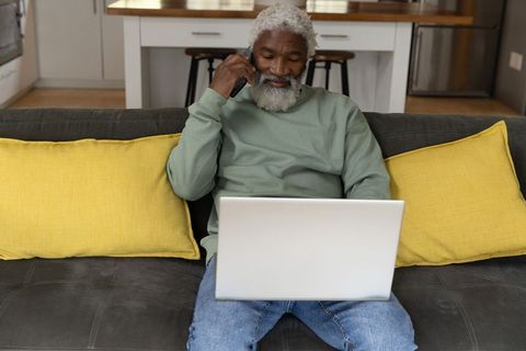 African american senior man talking on phone while working on laptop from sofa with yellow pillows