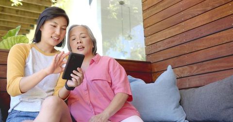 Adult daughter teaching older woman smartphone use while scrolling on sunlit patio bench