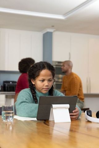 Multiracial girl using tablet at kitchen table while parents chatting in background
