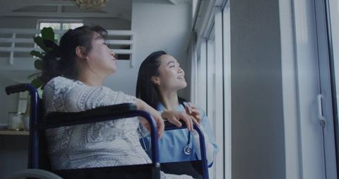 Compassionate caregiver in blue scrubs assisting wheelchair resident gazing out sunny window