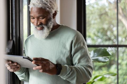 Mature african american man smiling while using tablet by sunlit window with potted plants