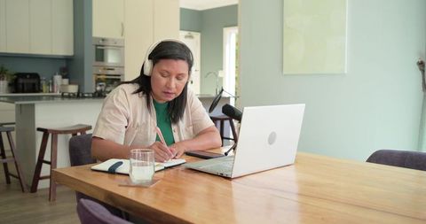 Mature woman wearing headset writing in notebook at home with laptop microphone for podcast