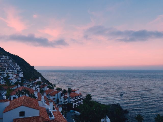 This image depicts a coastal village at sunset with a pink twilight sky over a calm sea. The picturesque Mediterranean-style houses with red rooftops complement the scenic view. Ideal for use in travel brochures, vacation planning websites, and promotional materials for scenic destinations. It captures tranquility and the beauty of coastal living.