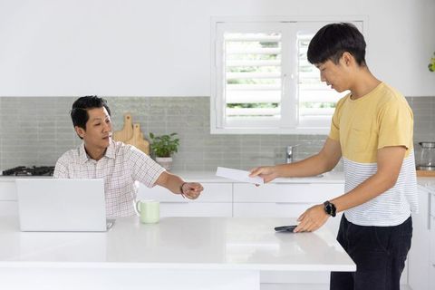 Asian men exchanging envelope at kitchen island while using laptop and smartphone