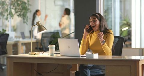 Excited Indian woman taking business call at modern coworking desk with laptop and coffee