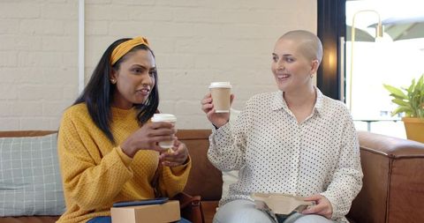 Diverse women sharing takeaway coffee and snacks, laughing on café couch
