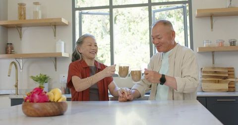 Senior asian couple sharing morning coffee and holding hands in bright contemporary kitchen