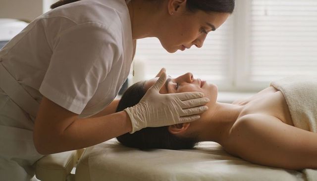Esthetician leaning over client wearing gloves in white uniform providing facial treatment