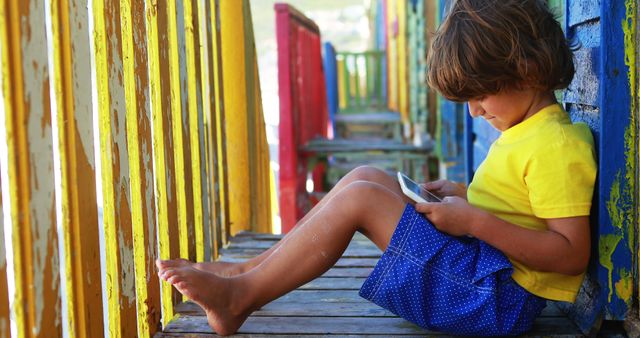 Young boy engrossed in playing with tablet while sitting on a colorful wooden balcony. Ideal for concepts of childhood, technology use, leisure during summer, and digital literacy. Can be used for educational materials, family lifestyle publications, or travel brochures highlighting family-friendly accommodations.