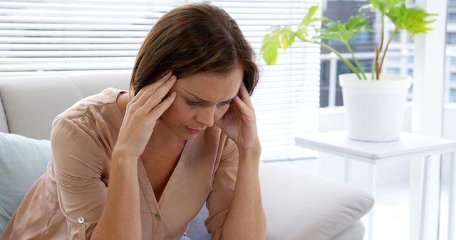 A woman sits in a modern living room holding her head with both hands, looking stressed or in pain. The environment is bright with natural light streaming in, and there is a potted plant beside her. This image could be used for articles or advertisements dealing with mental health awareness, stress management, healthcare services, work-life balance, or lifestyle blogs focusing on health and well-being.