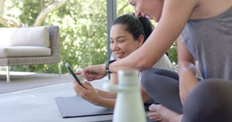 Women sharing fitness app on smartphone while reclining on yoga mat outdoors in sunlit patio