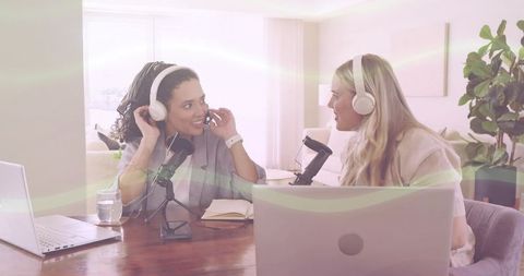 Two women recording podcast at home table wearing headphones and speaking into microphones