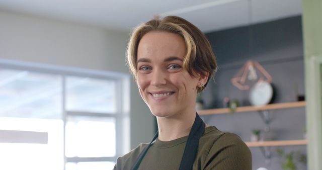 Young woman wearing casual clothing and apron smiling confidently in a modern kitchen setting. Natural lighting enhances the cheerful and relaxed atmosphere. Perfect for use in articles and advertisements related to cooking, lifestyle, home improvement, and self-confidence.