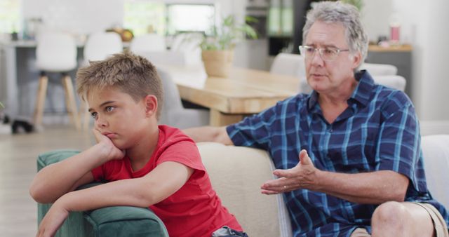 Elderly man with gray hair and glasses supports and talks to a sad boy wearing a red shirt while sitting on a couch in a bright, modern living room. Suitable for concepts of family care, empathy, multigenerational relationships, and emotional support.
