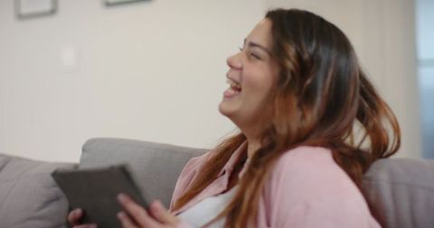 Laughing woman relaxing on gray couch holding tablet cozy home leisure digital lifestyle